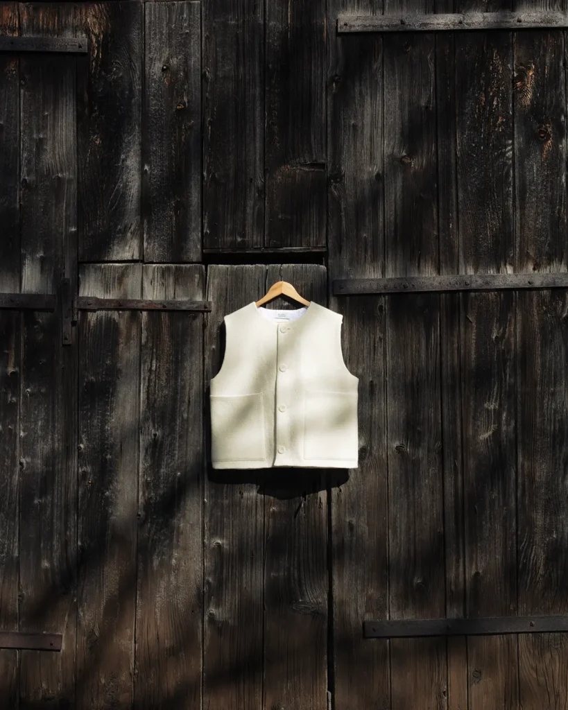 Photographie du Gilet d'Alpage de la marque Maison Douillet. Le gilet blanc est photographié contre la porte en bois d'une ancienne grange. Cette photographie nous rappelle que ce vêtement est inspiré d'un gilet de montagne, de la vie montagnarde et du patrimoine Alpin.